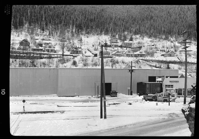 Photo of an unidentified building that has a train car near it, and a truck parked outside. The side of the Conoco gas station is visible. Houses are visible on the hill behind the building in question, and there is snow on the ground.