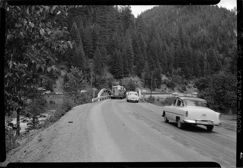 Photo of the Stansfield bridge on old highway 10 to Mullan. There is an oncoming truck crossing the bridge with several cars behind it, and two cars are seen driving the opposite direction. The area is surrounded by trees.