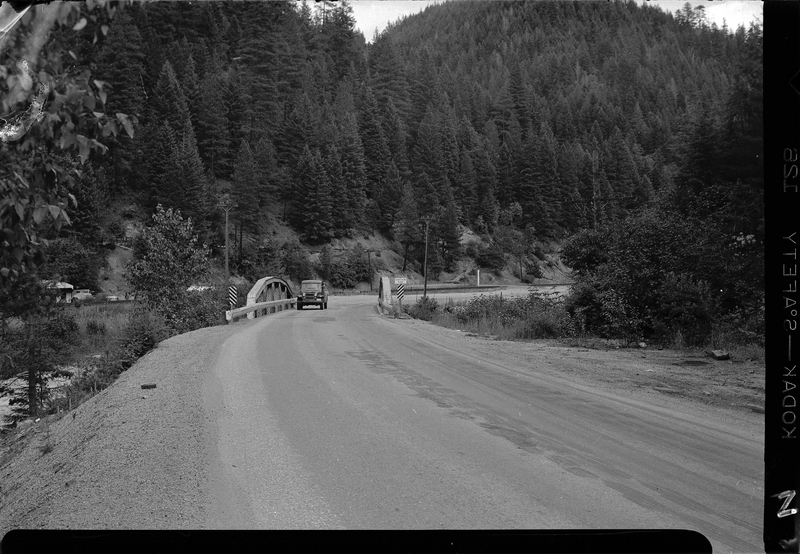 Photo of the Stansfield bridge on old highway 10 to Mullan. There is an oncoming car on the bridge. Trees surround the area.