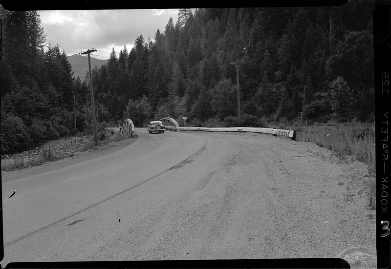 Photo of the Stansfield bridge on old highway 10 to Mullan. There is a car driving in the opposite direction from where the photographer is standing, driving over the bridge. The area is surrounded by trees.