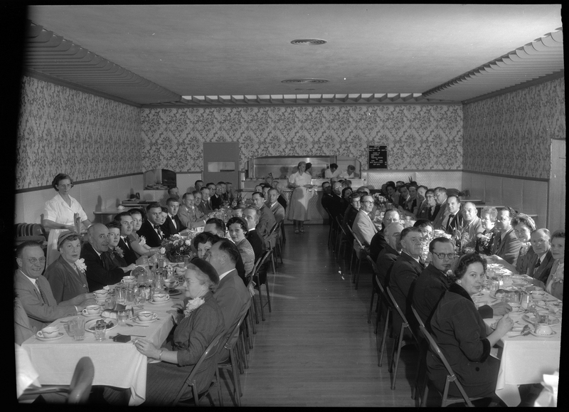Photo of the dining room inside the Benevolent and Protective Order of Elks building. There are two lines of tables on either side of the room that have men and women seated at them, and a few waitresses are handing out food. Most people sitting at the tables are looking at the camera for the photo.
