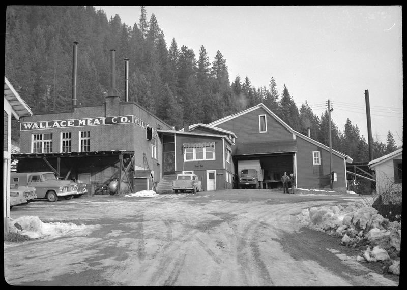Photo of the exterior of the Wallace Meat Company building. It is connected to what appears to be a house or office building, which has a garage attached. There are several cars parked outside and there is snow on the ground. There are also two men outside.