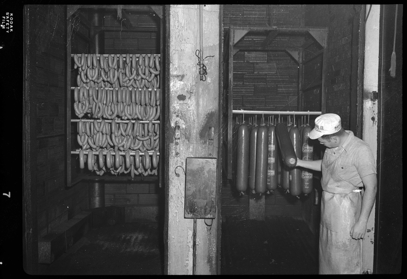 Photo of an unidentified man, probably a worker at the Wallace Meat Company, inspecting some meat that is hung up in one of two visible chambers in the building.