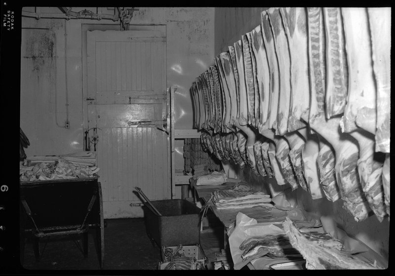 Photo of a line of unidentifiable animal carcasses hanging from meat racks inside the Wallace Meat Company building.
