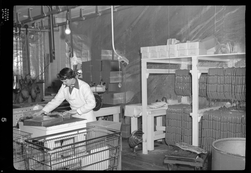 Photo of an unidentified woman working inside the Wallace Meat Company building. She is packing some meat at a workbench with a shopping cart next to her. There are meat racks full of sausages behind her.