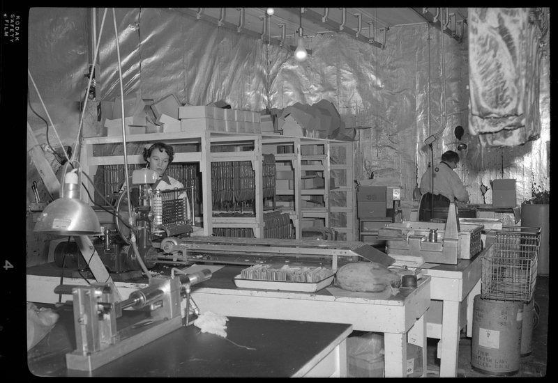 Photo of two unidentified women working inside the Wallace Meat Company building. They are working at separate workbenches, and one of them is operating some kind of machinery. There are meat racks with sausages hanging from them along the wall.