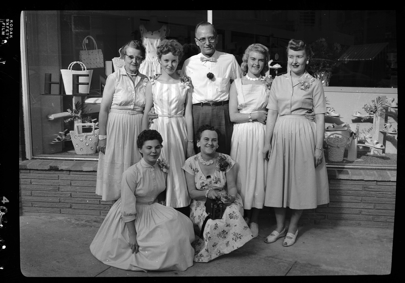 A man, probably Henry Kuhlruss, stands outside of a shop with six women. The women are all wearing nice dresses and everyone is smiling at the camera for the photo. They are probably standing outside of Henry Kuhlruss' clothes and shoe store for the grand opening.
