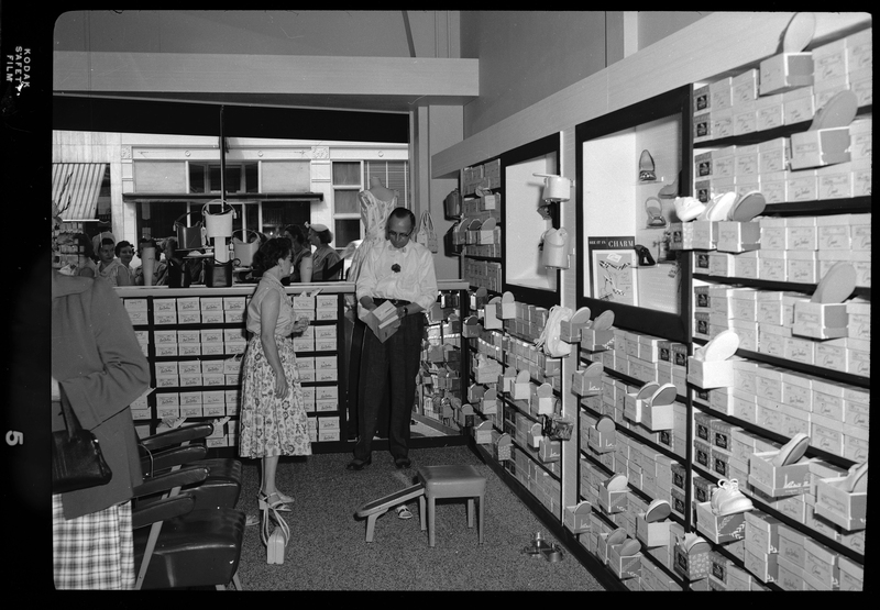 An unidentified woman is being helped by a man as she is shopping for shoes inside of Henry Kuhlruss' clothing and shoe store. A group of women can be seen outside the store through the window.