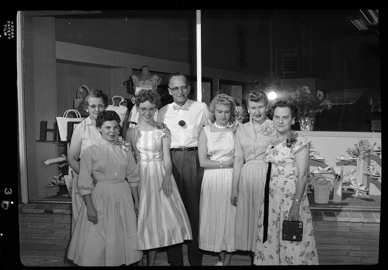 A man, probably Henry Kuhlruss, stands outside of a shop with six women. The women are all wearing nice dresses and everyone is smiling at the camera for the photo. 