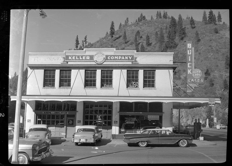 Photo of the Keller Company building in Wallace, Idaho. It is an authorized Buick dealership that has signs advertising used cars. There are several cars parked out front, and a gas pump on the side of the building.