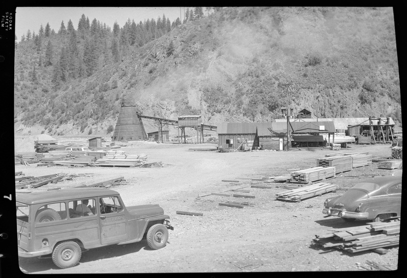 Photo of the Osburn Lumber Company lumber yard, pull of lumber piles. There is car and some machinery as well.