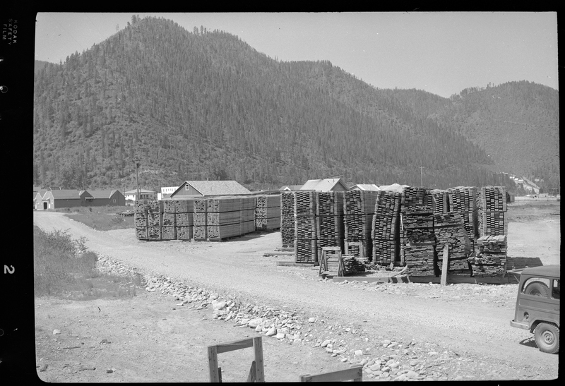 Photo of the Osburn Lumber Company lumber yard, pull of lumber piles. There is car partially in frame and some buildings in the distance as well.