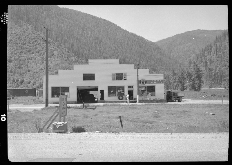 Photo of the Osburn Lumber Company building. There is a man standing outside and a garage door in the building that is open. There is a car inside the garage area and parked along the side of the building.