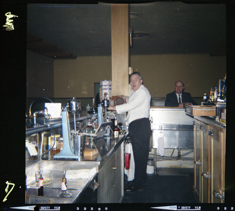 Color photo of two men inside Beauty Marte for its store opening. One man is working behind a counter and the other is sitting at the counter. They are both looking at the camera.