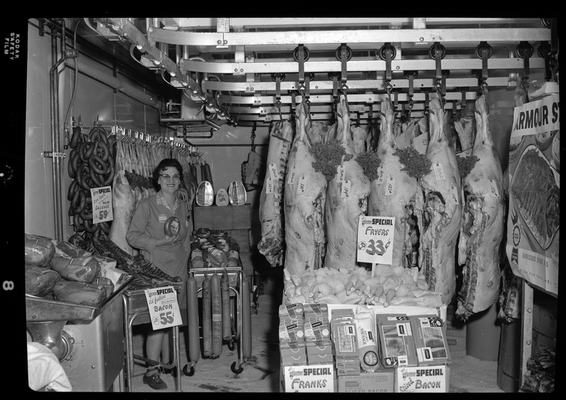 Photo of an unidentified woman standing with a meat display inside City Meat Market. Sausage is listed at $0.59 per pound and bacon is listed at $0.55 per pound.
