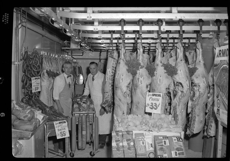 Photo of two unidentified men standing with a meat display inside City Meat Market. Sausage is listed at $0.59 per pound and bacon is listed at $0.55 per pound.