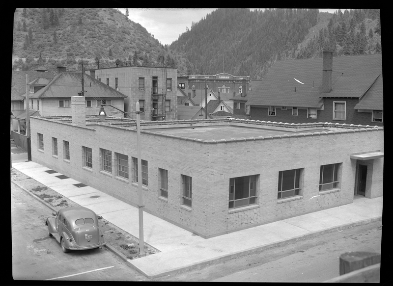 Photo of the "telephone building" on 4th Street and Bank Street in Wallace, Idaho built in the 1940's. The photo is taken across from one corner of the building and from above, showing two sides and the roof. There is a car parked on the side of the road next to it.