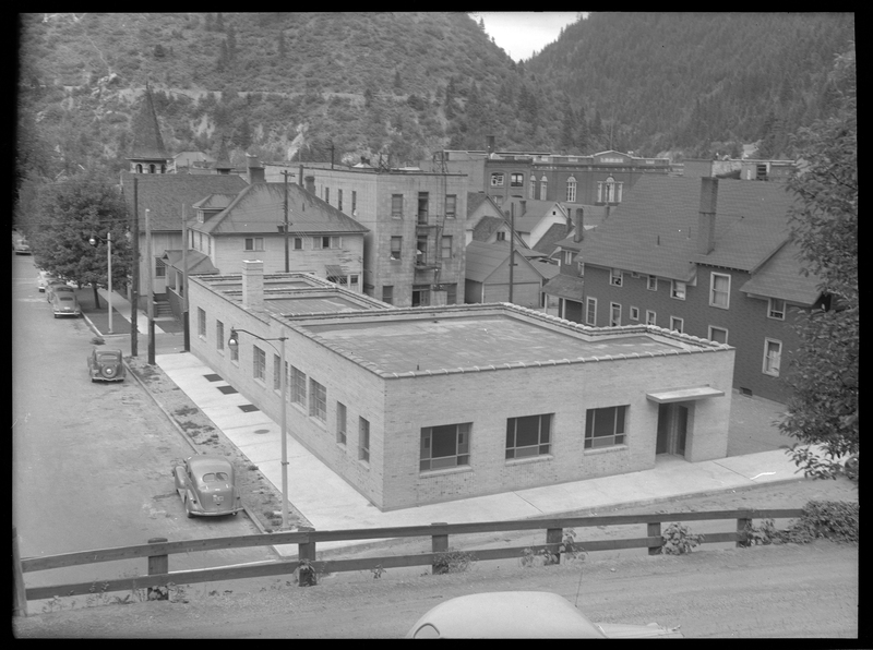 Photo of the "telephone building" on 4th Street and Bank Street in Wallace, Idaho built in the 1940's. The photo is taken across from one corner of the building and from above, showing two sides and the roof. There are a couple of cars parked on the side of the road next to it.