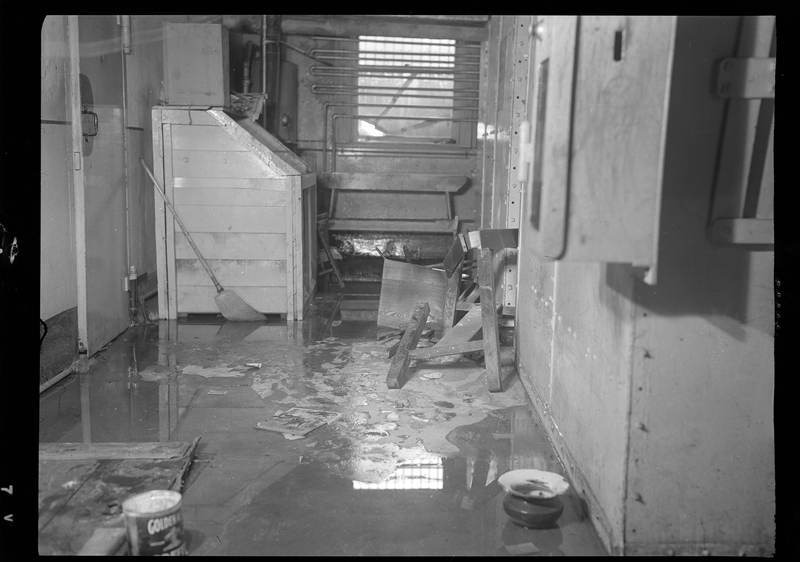 Photo of the inside of the Shoshone County Jail (in the basement of the Wallace Courthouse). There is a lot of water pooled on the floor and some of the furniture appears to be overturned.