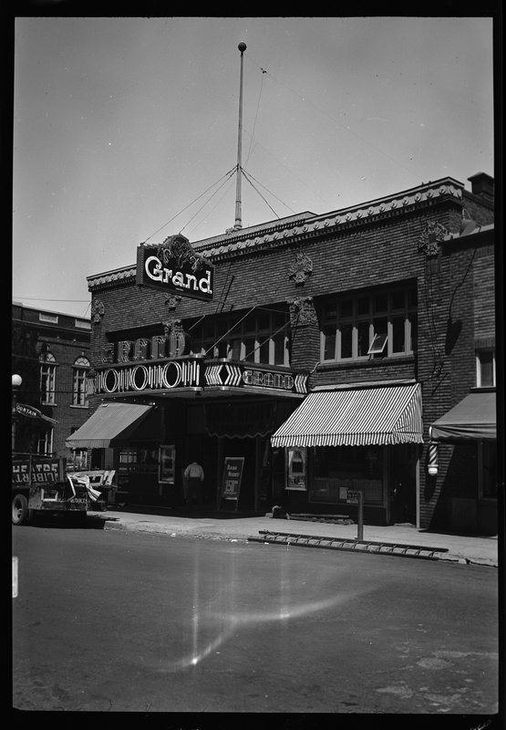 Front view of the Grand Theatre building. The building has an overhang with a "Grand" sign that appears to be neon, and awnings over the windows.