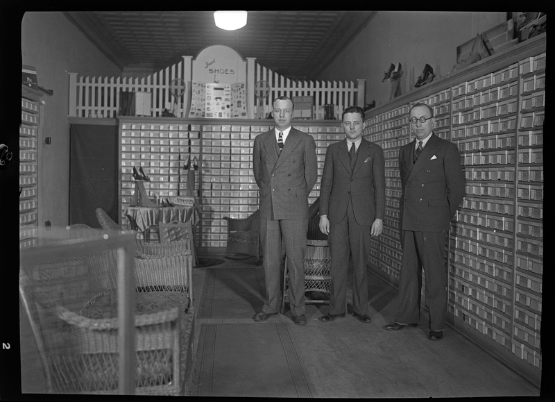 Photo of three men standing inside of Murphy's Bootery, later renamed Pullman Bootery. There are some chairs throughout the shop and the walls are lined with shelves of shoe boxes.