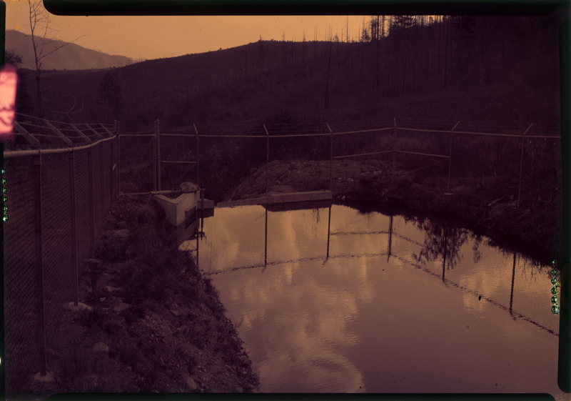 Color photo of a small body of water, possibly a controlled reservoir or canal, that is surrounded by a chain link fence.
