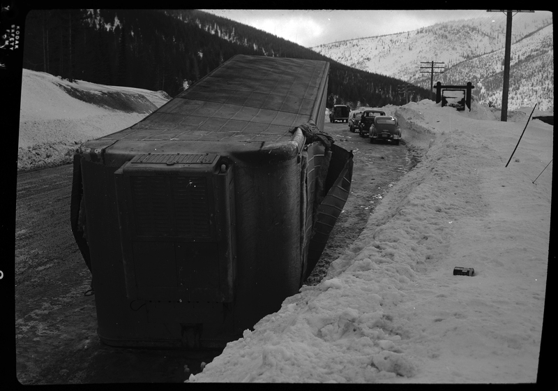 Photo of a tipped over trailer on the road. It's no longer attached to the cab of the semi truck and it shows signs of damage. There is snow on the ground and several cars parked on the side of the road further ahead.