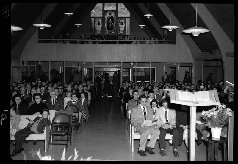 Photo of a Congregational Church gathering inside of a church. Men, women, and children are seated in the pews.