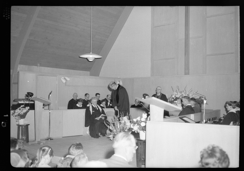 Photo of the pastor of the Congregational Church gathering about to address the crowd. There are a few people sitting on stage around him, possibly singers.