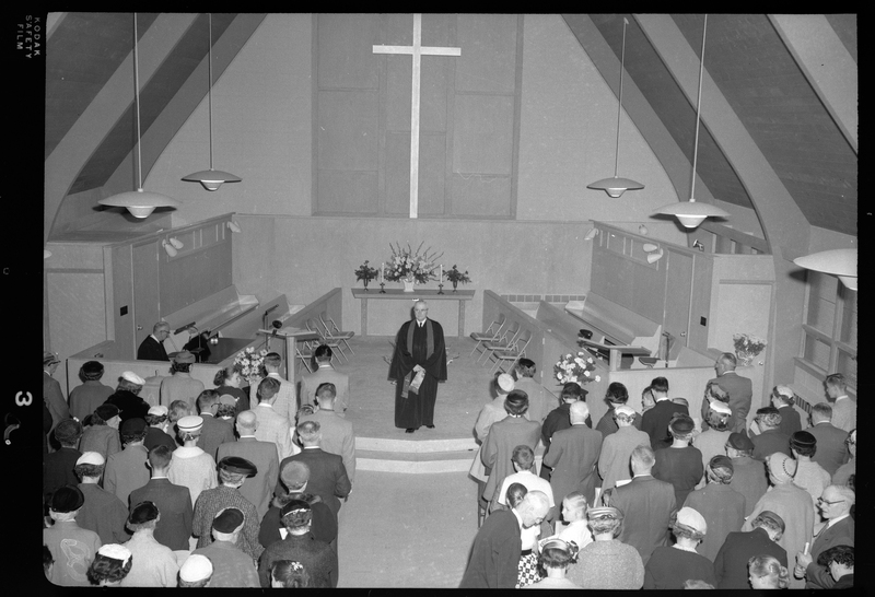Photo of the pastor of the Congregational Church gathering addressing the crowd of men, women, and children. There is another man on the stage sitting behind a piano.