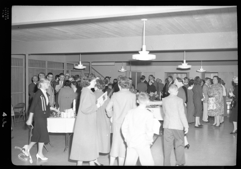 A group of men and women are gathered together for a Congregational church gathering. There are a few tables in the room that appear to have food on them, with people standing all around the room.