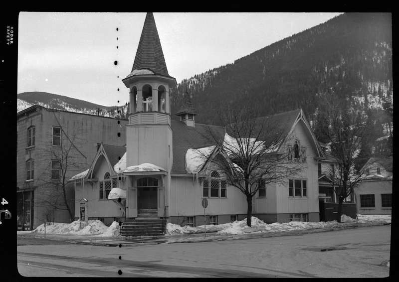 Photo of a church building on a street corner. There are a few trees along the sidewalk, and there is melting snow on the roof and ground of the church. The negative has some minor damage on it.