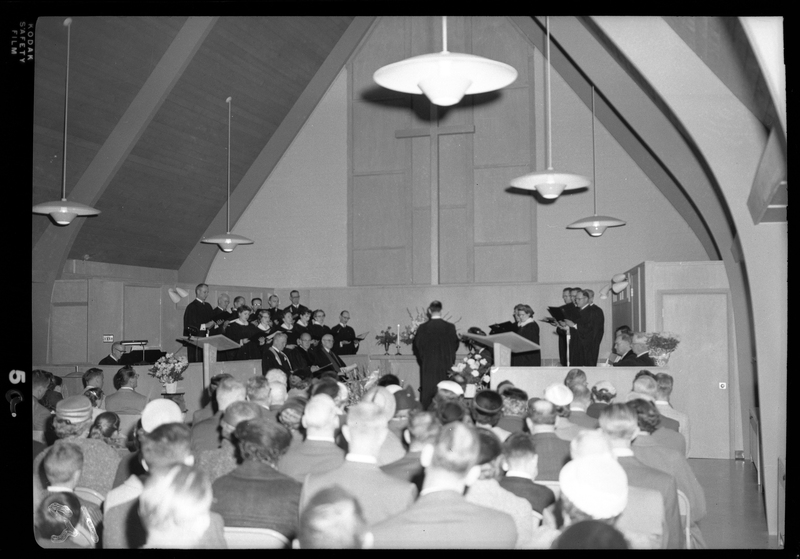 Photo of a Congregational Church gathering inside of a church. Men, women, and children are seated in the pews and there are people standing on the stage, possibly the choir.