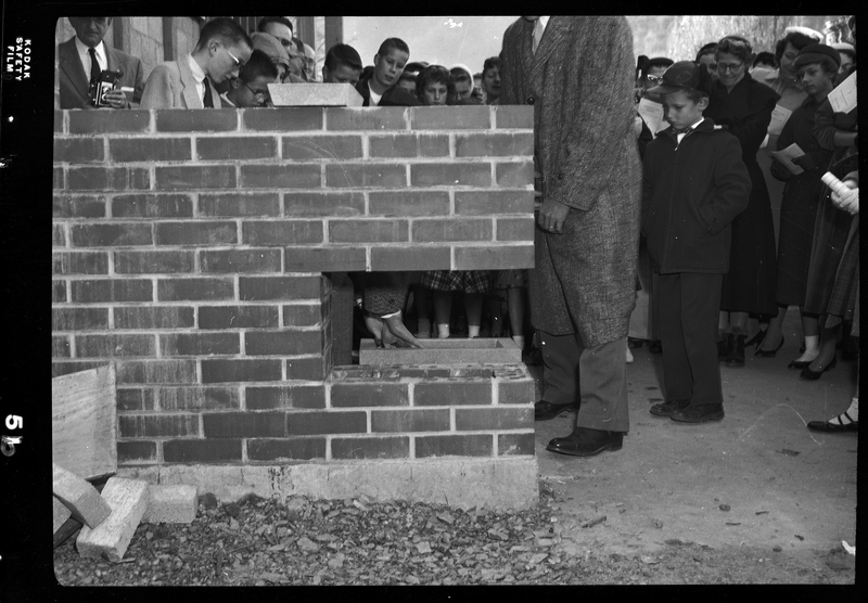 Photo of an unidentified building under construction. There is a cut out section of a brick wall and a cinder block next to it to be placed in the opening. A small crowd of men, women, and children gather around as two men prepare to place the cinderblock.