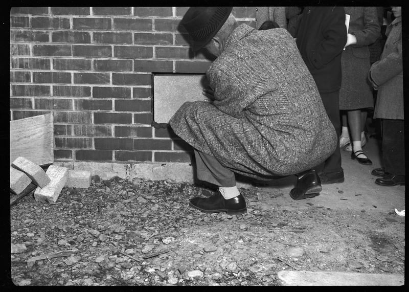 Photo of an unidentified man placing a cinder block into a cut out section of a brick wall while people stand around and observe.