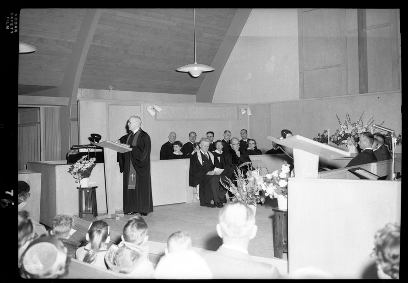 Photo of a Congregational Church gathering inside of a church. Men, women, and children are seated in the pews and there are people seated on stage, possibly the choir, and a man is addressing the crowd.
