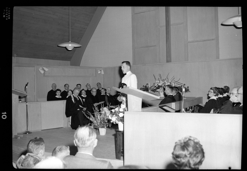 Photo of a Congregational Church gathering inside of a church. Men, women, and children are seated in the pews and there are people seated on stage, possibly the choir, and a man is addressing the crowd.