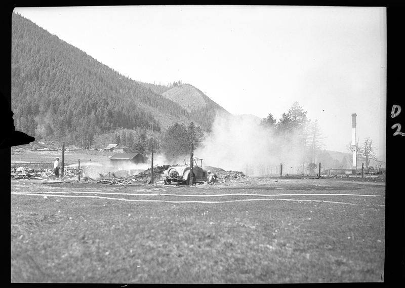 Photo of a man who appears to be spraying water on what appears to be a fire in an open field. There is a car parked on the side of the road.