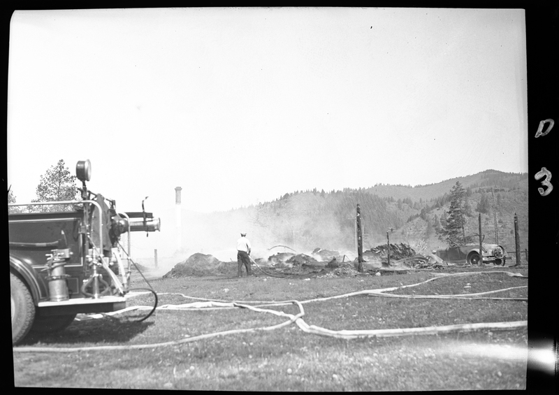 Photo of a man who appears to be spraying water on what appears to be a fire in an open field. There is a car parked on the side of the road.