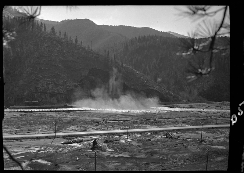 Photo of an unidentified outdoor scene. The area is muddy and empty, and there is a river in the background. There is a cloud of dust that appears to have originated on the opposite side of the river.