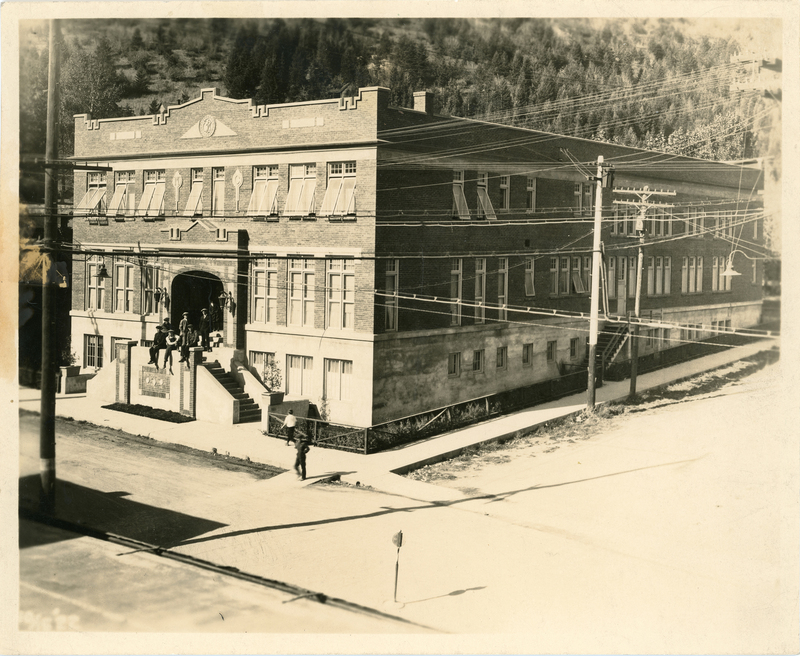 Photo of the Morning Club building in Mullan, Idaho. There are several men standing and sitting on the steps that lead to the entrance of the building, and a few people walking on the street next to the building.