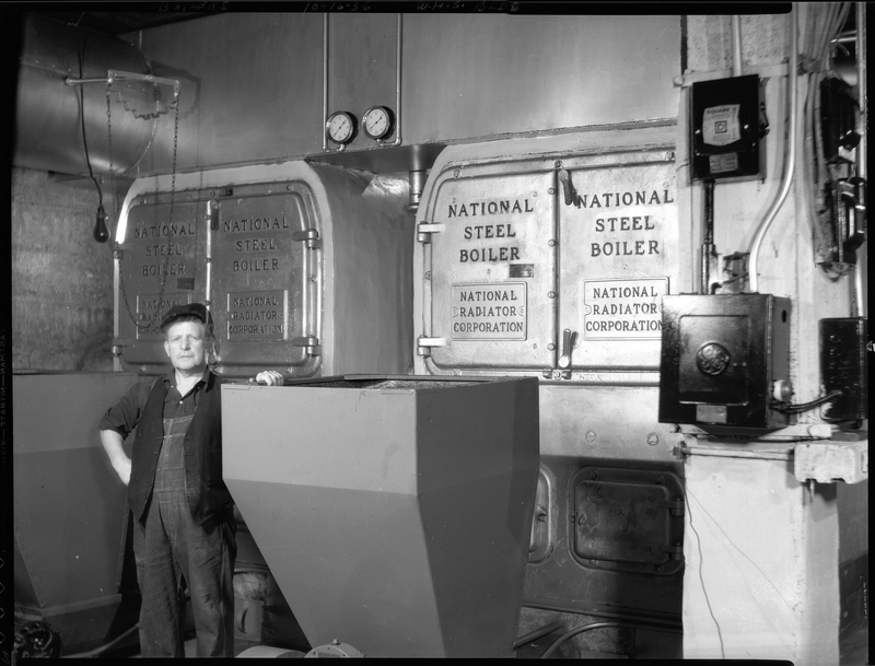 Photo of an unidentified man standing next to the National Steel Boilers that are inside of the Wallace High School building. There are signs on the boilers that read, "National Steel Boiler; National Radiator Corporation."