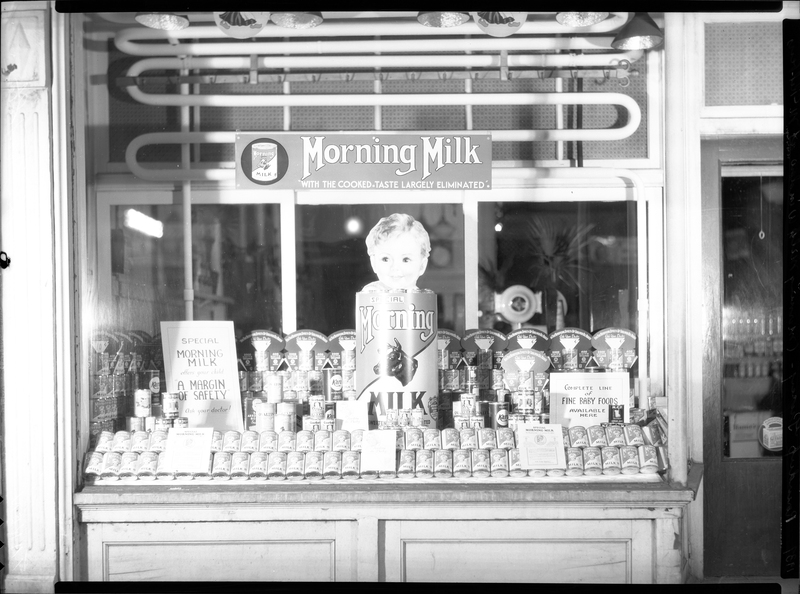 Photo of the Morning Milk window display inside of Roundup Grocery. There are three signs, one that reads, "Morning Milk; with the cooked taste largely eliminated," another that reads, "Special Morning Milk; A margin of safety; ask your doctor," and the last one reads, "Complete line of fine baby foods available here."