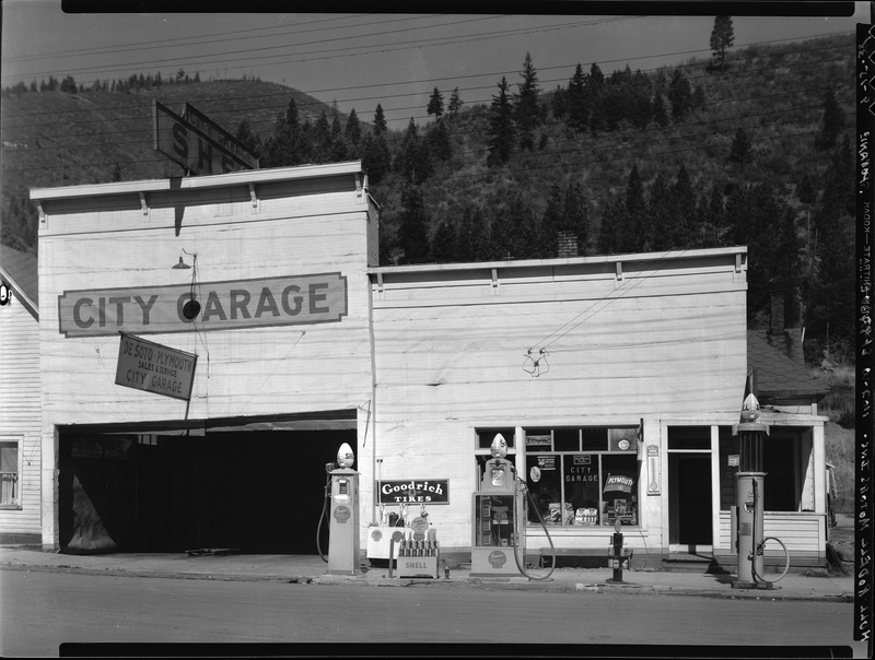 Photo of the exterior of the City Garage. There are several signs on the outside advertising products and services, as well as three gas pumps. The garage door is open.