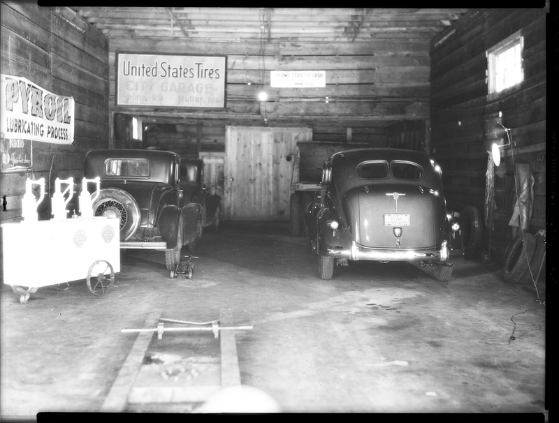 Photo of four vehicles parked inside of the City Garage building. There are three cars and a truck, and there is a sign on the back wall that reads, "United States Tires; City Garage; Phone - 99; Mullan, Idaho."