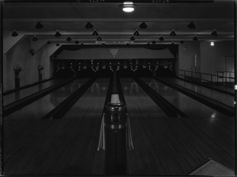 Photo of the Wallace Bowling Alley lanes looking towards where the pins are. There is a sign on the wall over the lanes that reads, "Please observe the foul line." The photo is very dark.