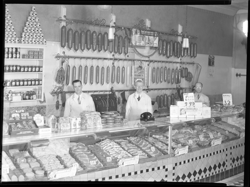Three unidentified men stand behind the display counter at City Meat Market. There are sausages hanging from meat racks on the wall behind them, and raw meat in the display cases.