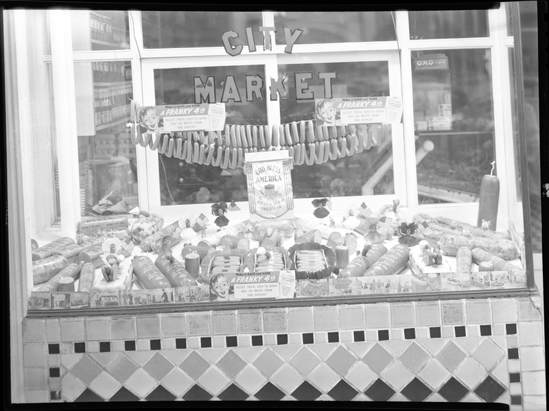 Photo of the window display at City Meat Market. There are various cured meats displayed in the window, and several signs that read, "Make it safe and sane; a frankly 4th; select these easy-to-serve July 4th meats from this display!"