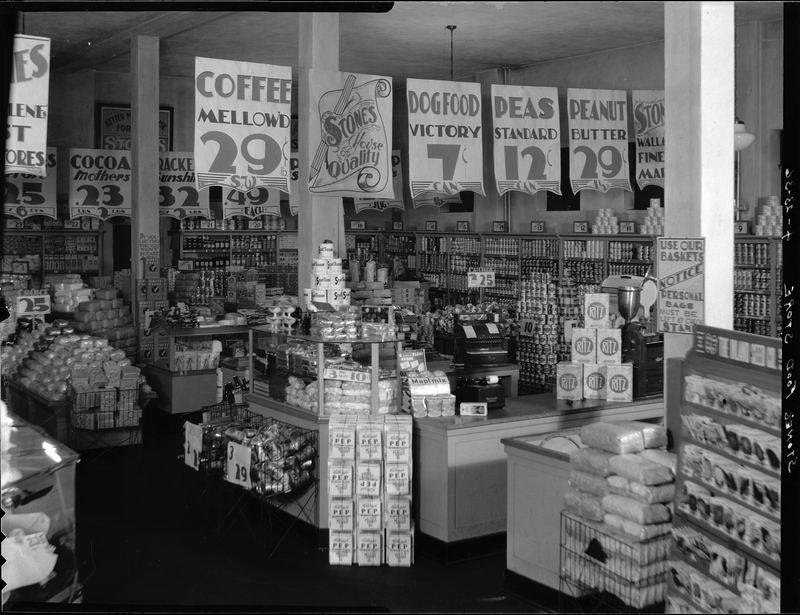 Photo of the interior of Stone's Food Stores. There is various foodstuffs lining the walls and shelves, and signs advertising products throughout.