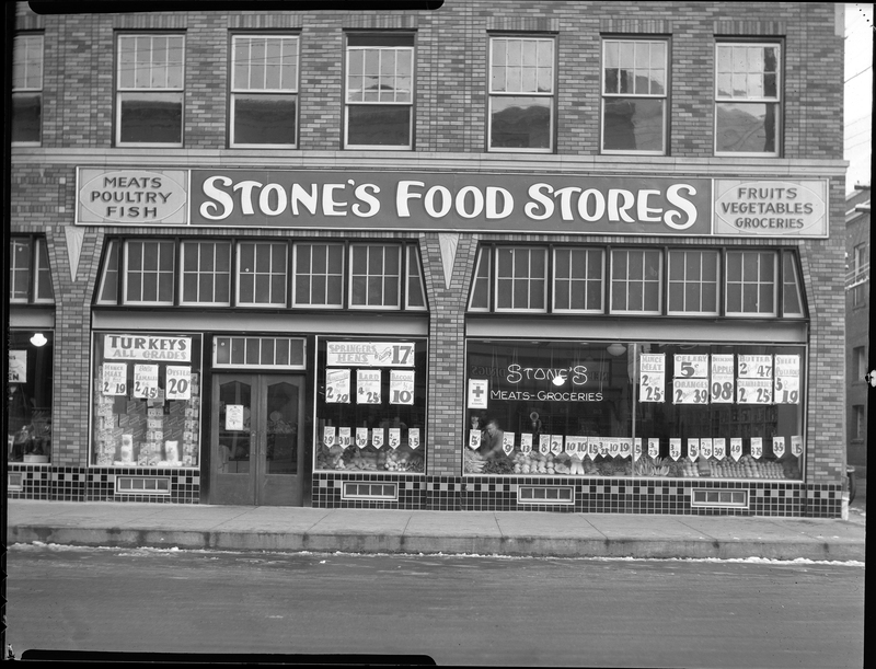 Photo of the storefront of Stone's Food Stores. There are several signs in the windows advertising various food products, and produce is arranged in the window displays.